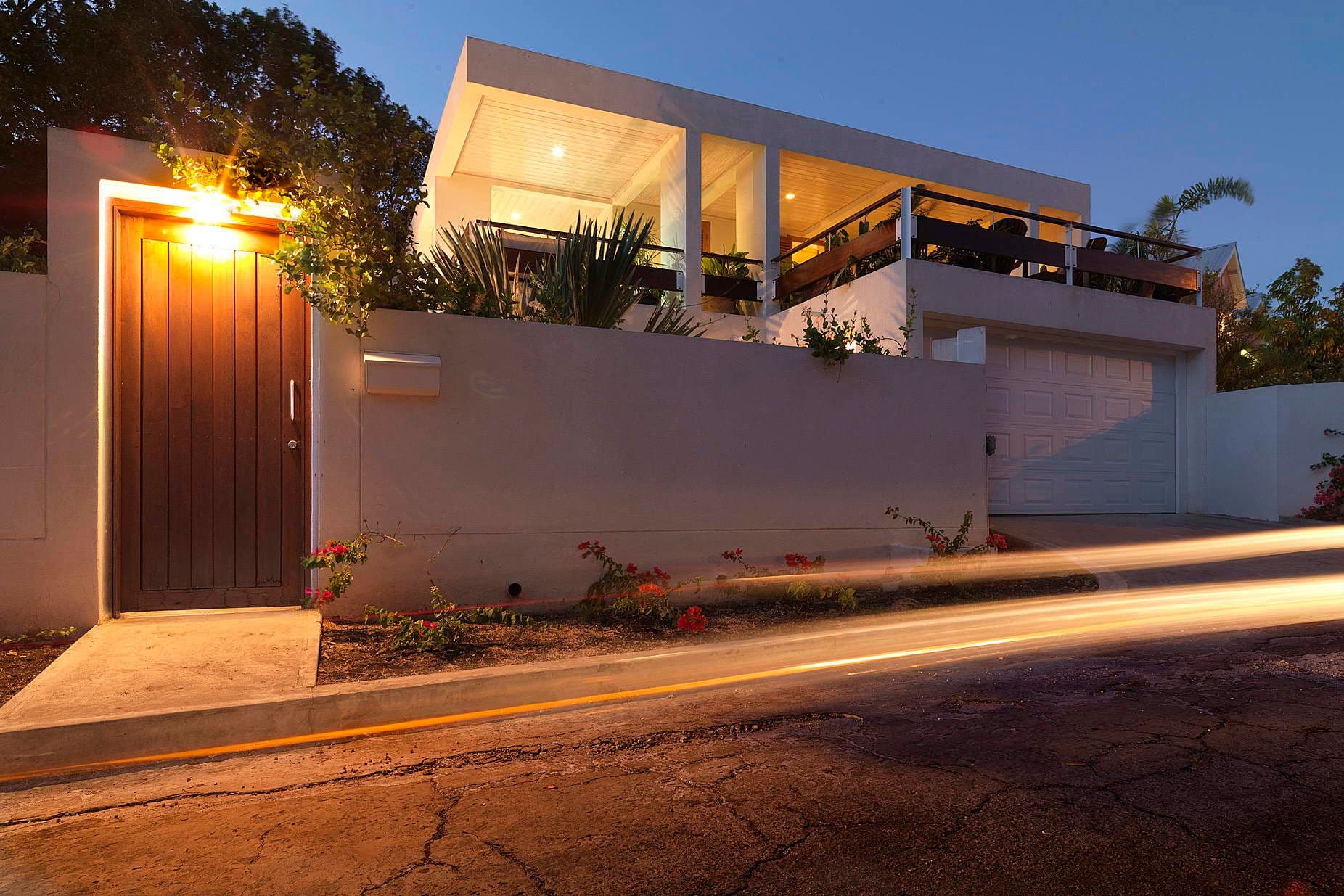 Modern white house at night with a lighted entrance gate and street light trails.