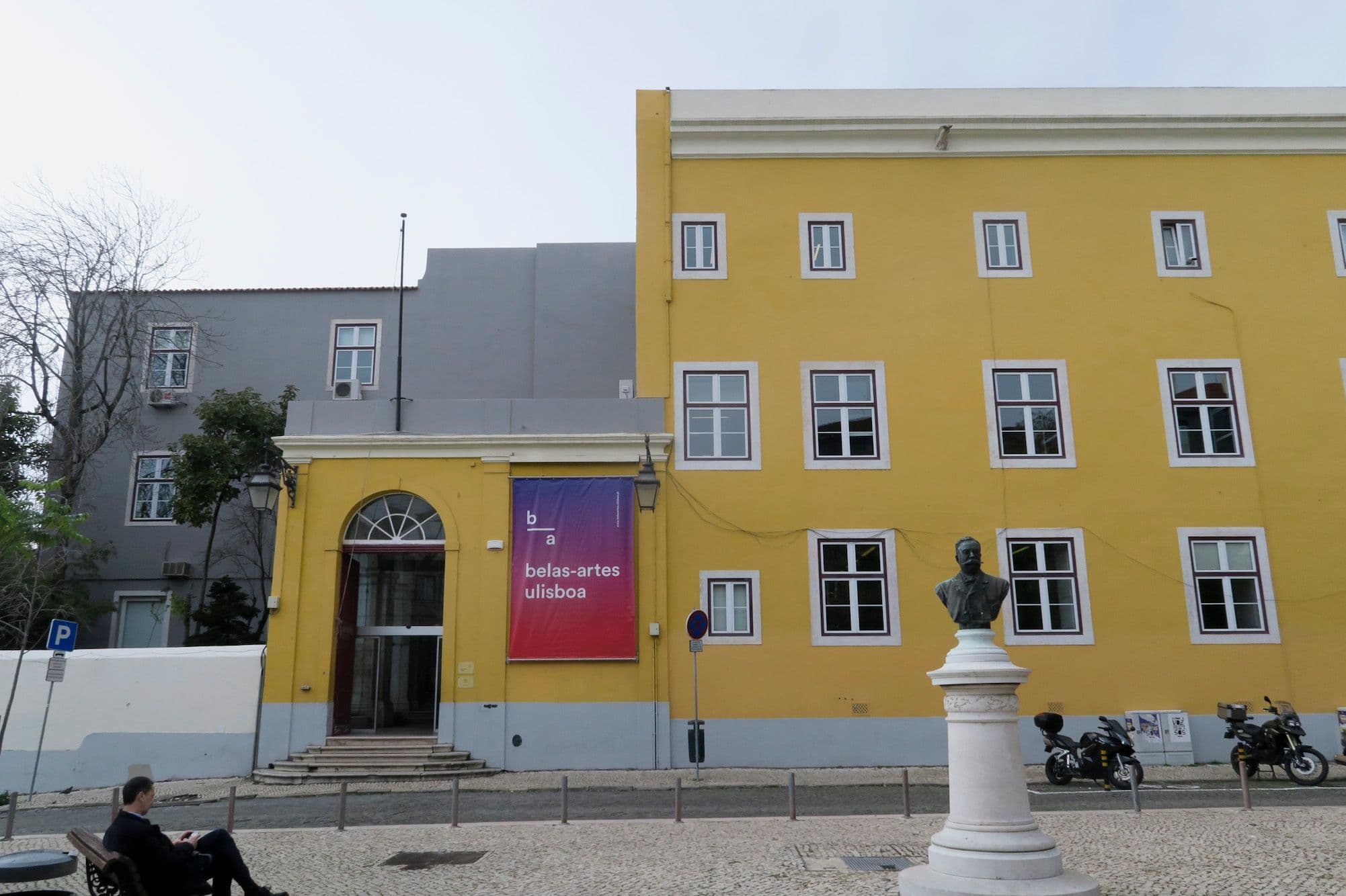 A bright yellow building with a bust statue in front, next to a grey building.