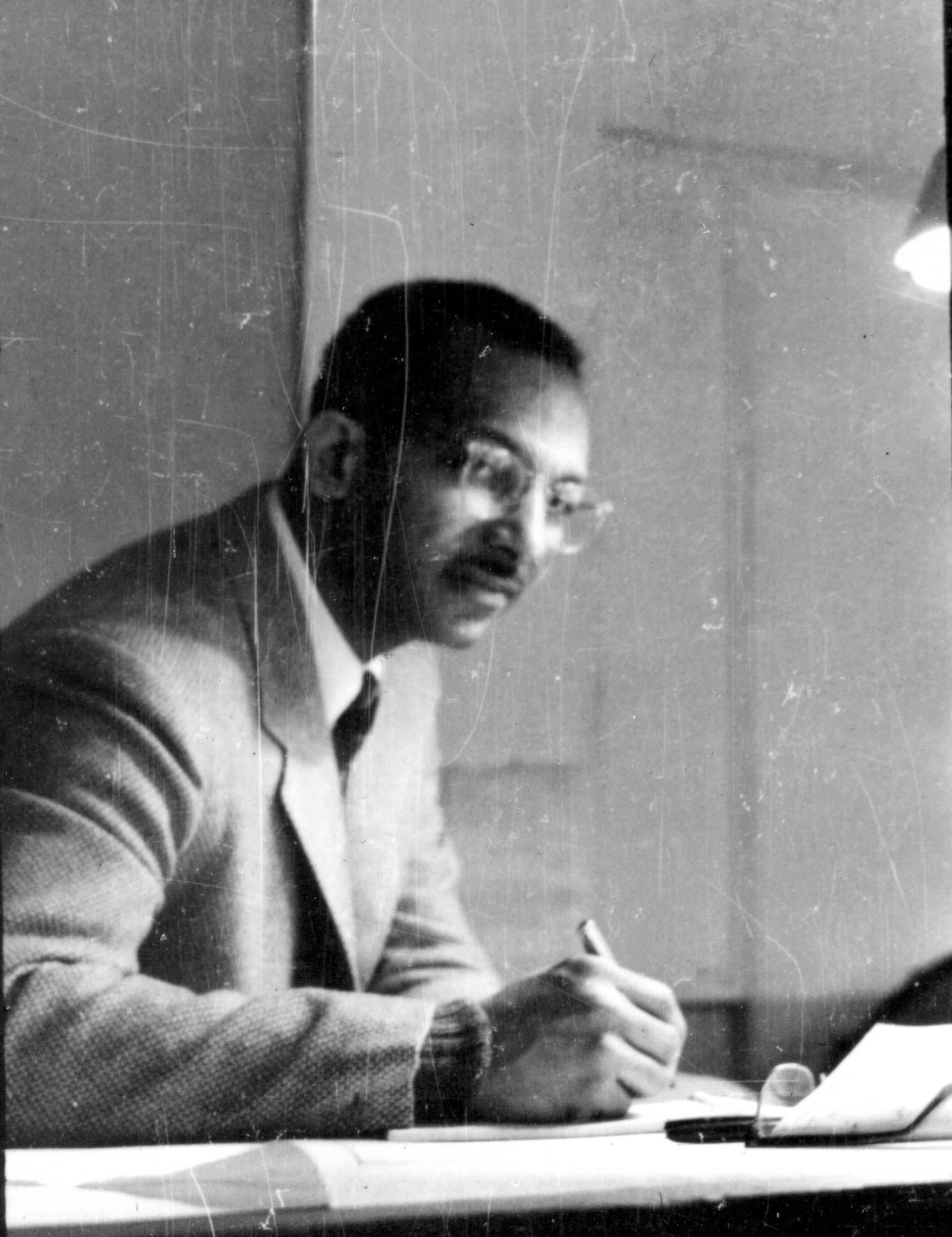 Man in glasses and a suit writing at a desk, illuminated by an overhead light.