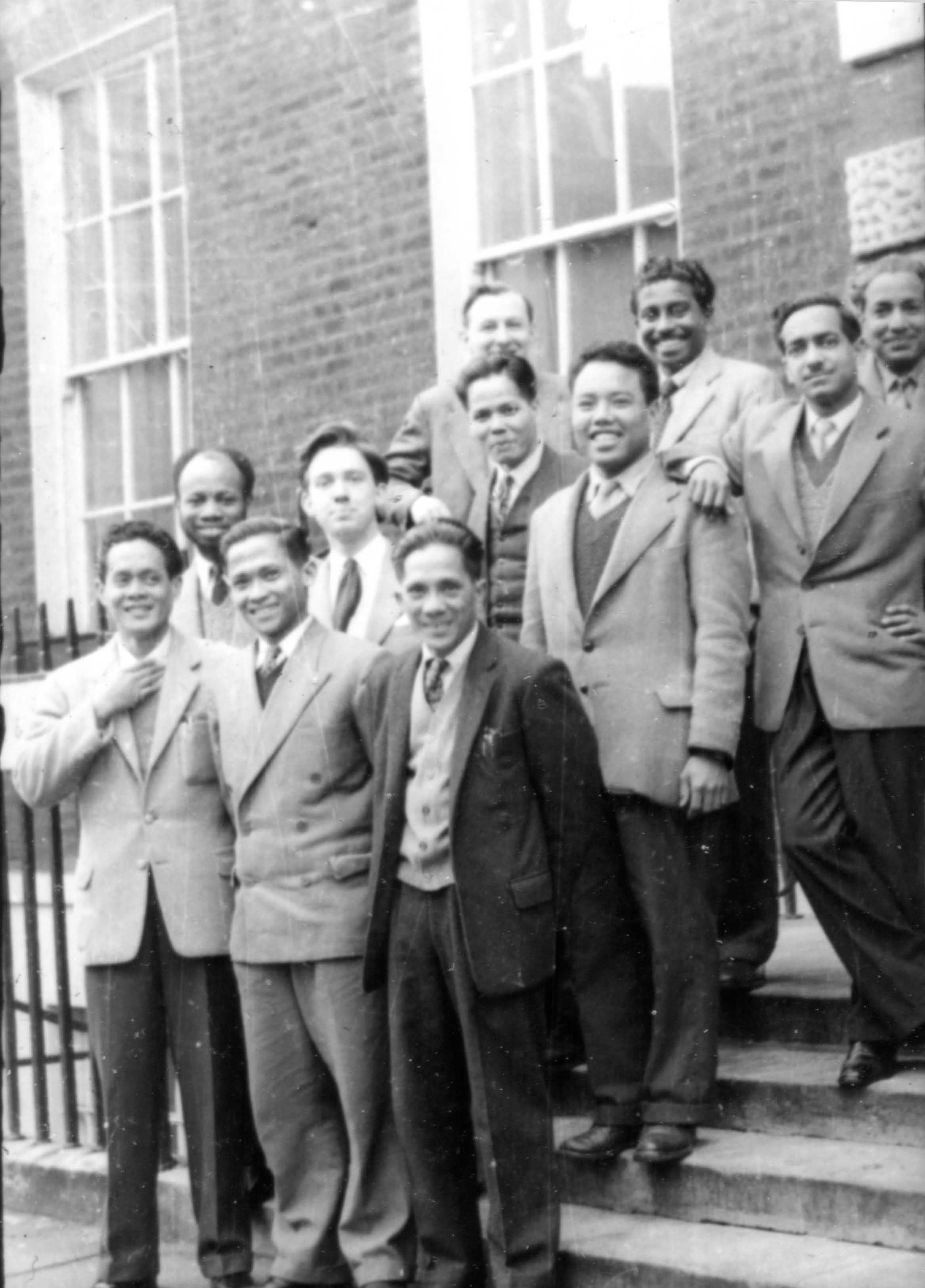Group of ten men in suits and ties posing on the steps of a brick building.