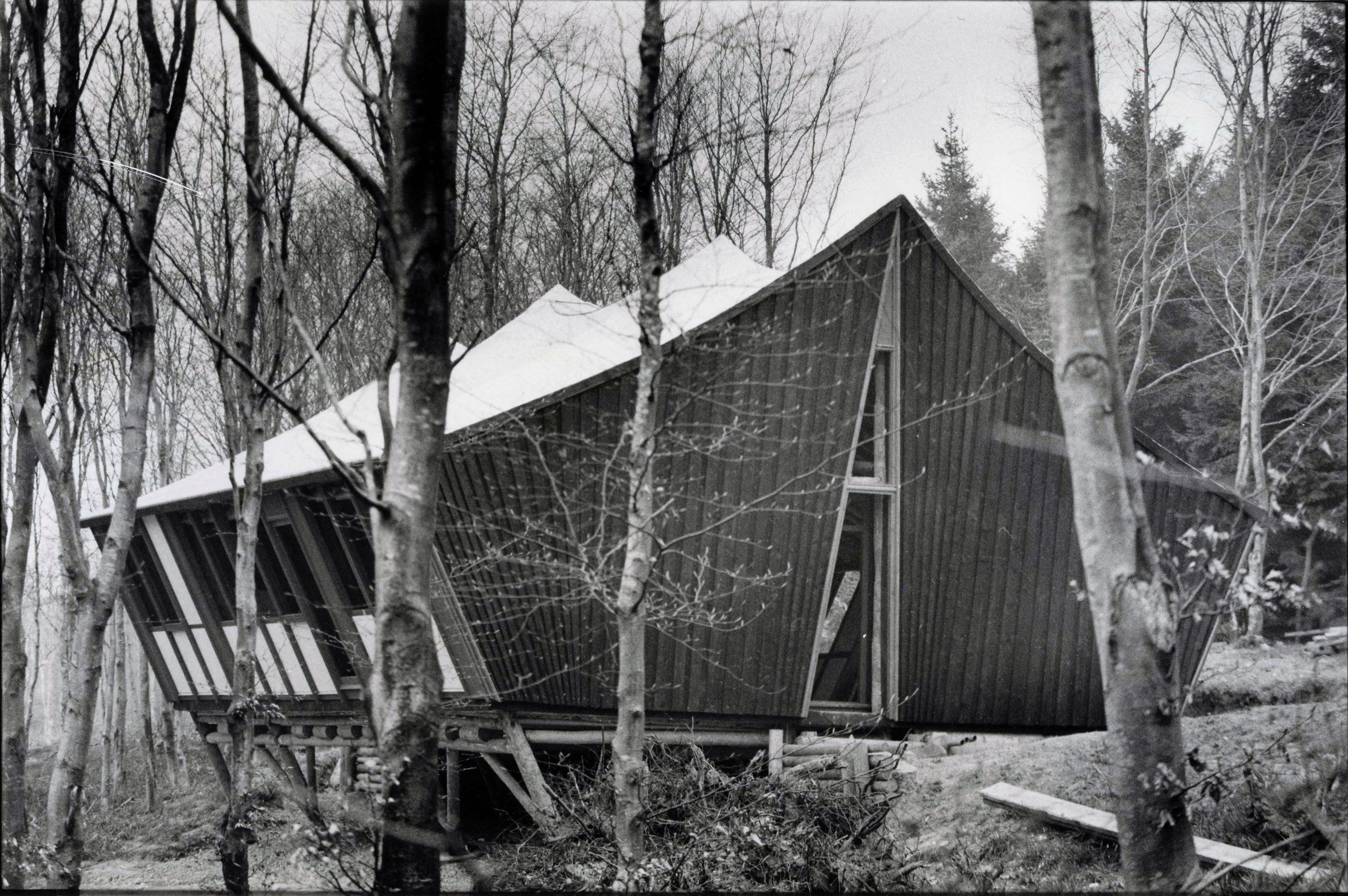 A black and white photo of a timber building with a pitched roof and an A-shaped door sitting among bare tree trunks in winter.